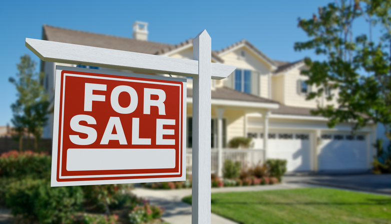 A close-up of a white wooden "For Sale" sign with a bright red background, standing in the front yard of a large, two-story suburban house.